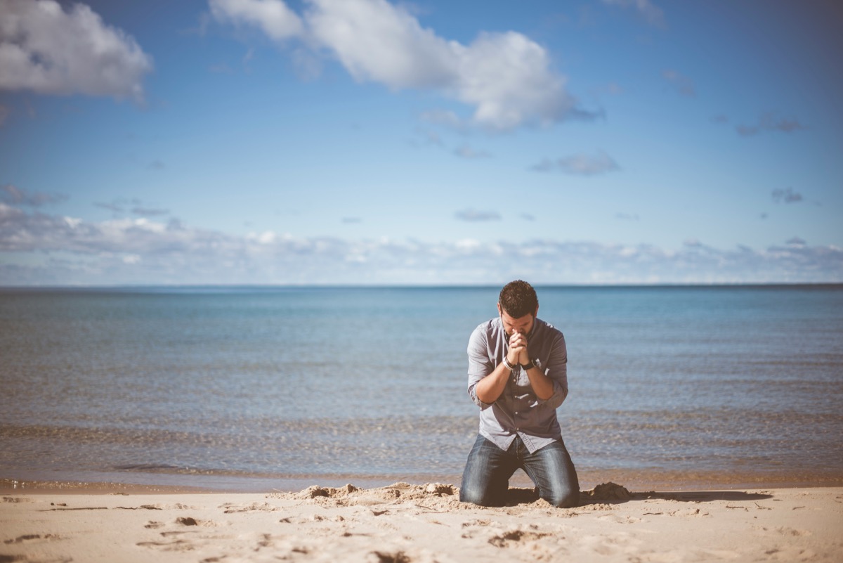 A man kneeling in prayer on a beach.