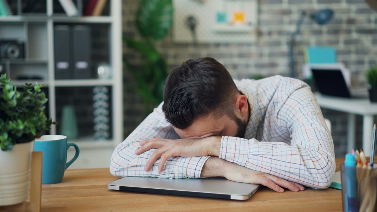 A tired man resting his head on his arms at a desk.